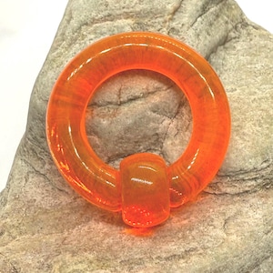 May include: An orange, circular body jewelry piece with a cylindrical bar. The transparent piece is set against a gray stone background. The jewelry is a vibrant, eye-catching accessory.