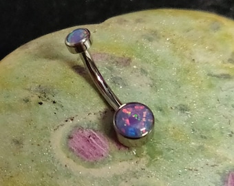 Peut inclure: Un piercing de nombril incurvé avec une barre argentée et deux pierres d'opale rondes et irisées. Les opales présentent des nuances de bleu, de violet et de rose. Le bijou est posé sur un fond texturé multicolore.