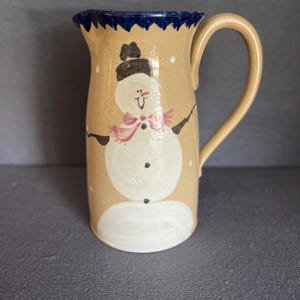 May include: A tan ceramic pitcher with a blue rim and a hand-painted snowman design. The snowman has a black hat, a pink scarf, and black eyes and buttons.