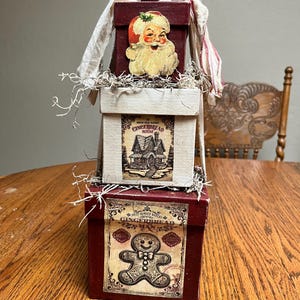 May include: A three-tiered stack of red and white boxes decorated with vintage Christmas imagery. The top box features a Santa Claus illustration, the middle box has a gingerbread house illustration, and the bottom box has a gingerbread man illustration. The boxes are adorned with festive details like holly and ribbons.