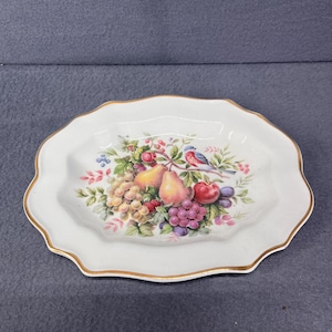 May include: A white ceramic plate with a scalloped edge and a gold trim. The plate features a colorful fruit and bird design with pears, grapes, and a bluebird.