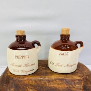 May include: A pair of ceramic salt and pepper shakers in a jug shape. The shakers are brown and cream-colored with cork stoppers. The pepper shaker reads "Pepper, Almost Heaven, West Virginia." The salt shaker reads "Salt, Old Fort Niagara."