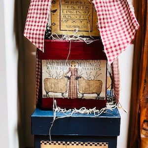 May include: Three stacked wooden boxes with a red and white gingham fabric bow on top. The top box has a vintage alphabet design with the text "ELIZABETH 1781". The middle box has a vintage illustration of a woman with two sheep and the text "SHE SEWS WELL AND I LAA AND WORKS WILLINGLY WITH HER HANDS". The bottom box has a vintage illustration of a raven and a pepper and the text "SIMPLY PRIMITIVE".