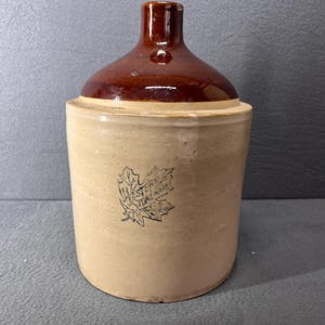 May include: A brown and tan stoneware crock with a brown glazed top. The crock has a maple leaf design with the text "WESTERN STONEWARE" on it.