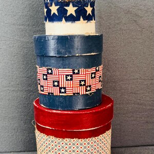 May include: Three round, stackable boxes with red, blue, and white fabric trim. The top box is white with a blue and white star pattern, the middle box is blue with a red and white striped pattern, and the bottom box is red with a white and blue diamond pattern.
