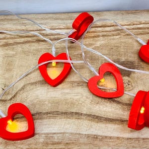 May include: A string of red heart-shaped lights with a warm yellow glow. The lights are strung on a clear wire and are arranged on a wooden surface. The heart lights are a festive decoration.