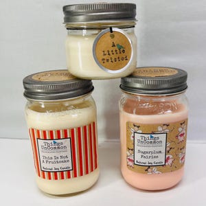 May include: Three glass jar candles with silver lids. One candle has a tag that reads "A Little Twisted." Another has a label that says "This Is Not A Fruitcake." The third candle is labeled "Sugarplum Fairies."