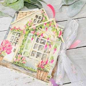 May include: A watercolor-style journal featuring a cottage scene with pink flowers and green vines. The journal has a white ribbon tie and pink accents. Wicker baskets with pink flowers are in the foreground.