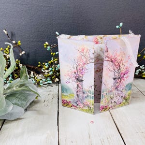 May include: A decorative box with a floral design featuring a tree with pink blossoms, a white fence, and a stone pillar. The box is open, revealing the interior, and has white lace trim. The background is a soft, pastel color palette.