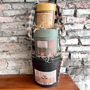 May include: Three stacked round boxes with a plaid fabric draped over the top. The top box is yellow with black lettering and a bird design. The middle box is green with a brown label that says "Never Enough Rhyme". The bottom box is black with a brown label that says "Sheers Wool and Flax and Works Willingly With Her Hands".