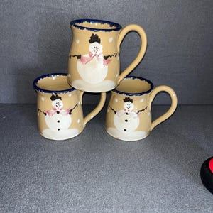 May include: Set of three ceramic mugs with snowman designs. Each mug is beige with white snowmen wearing pink scarves and black hats. The mugs have blue rims and white polka dots.