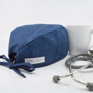 May include: A navy blue surgical scrub cap with a paisley pattern. The cap has a tie in the back and a small white label with text. A white mug and a stethoscope are next to the cap.