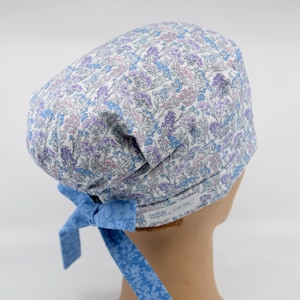 May include: A floral patterned scrub cap in shades of white, blue, and purple. The cap features a light blue bow at the back and a tag with text. The fabric is covered in a delicate floral print.