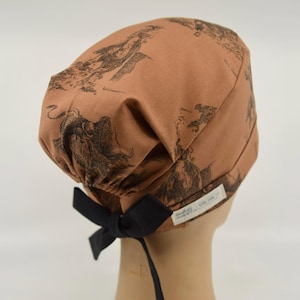 May include: A tan scrub cap featuring a dark brown illustrative print of rodeo cowboys and bulls. The cap has an elasticized back with a solid black ribbon tie and a small white fabric label stitched to the side with black text.