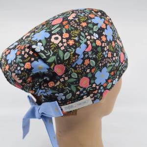 May include: A floral patterned scrub cap featuring a black background with pink, blue, and orange flowers and green leaves. The cap has a light blue tie with gold stars. The cap is designed to be worn on the head.