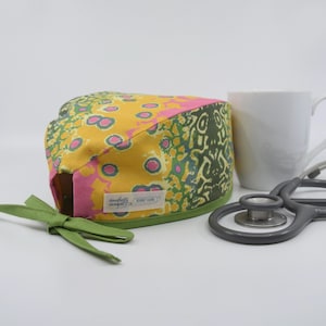 May include: A patterned scrub cap with a green tie, featuring yellow, pink, and green designs. A white mug and a stethoscope are in the background. The cap has a label that reads "daugherty designs".