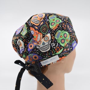 May include: A black surgical scrub cap featuring a colorful Day of the Dead sugar skull pattern in orange, green, purple, and white. The cap has a black tie with gold stars and a small white tag that reads "daugherty designs".