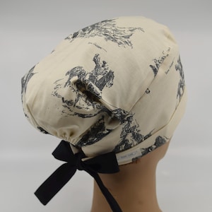 May include: Cream-colored scrub cap featuring a black and white toile print of riders on horseback. The cap has a black bow at the back for adjusting the fit. A small tag with text is visible on the side.