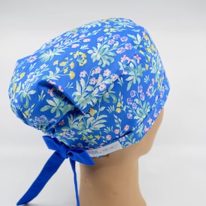 May include: A blue scrub cap featuring a repeating floral pattern in shades of yellow, light purple, and green. The cap includes a solid blue fabric tie at the back for an adjustable fit and a small white rectangular label on the side band.