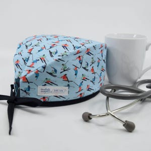 May include: A light blue surgical scrub cap with a pattern of skiers in various colors. The cap has a black tie closure. A white mug and a stethoscope are in the background.