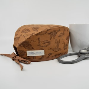 May include: A brown scrub cap featuring a repeating pattern of duck illustrations. The cap has a tie closure and a small label with text. A white mug and a stethoscope are in the background.