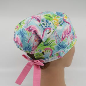 May include: A scrub cap featuring a vibrant pattern of pink flamingos, green palm leaves, and blue foliage on a light blue background. The cap has a gathered back with two solid pink fabric ties and a small white rectangular label on the side.