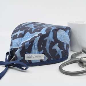 May include: A navy blue surgical scrub cap featuring a pattern of blue whale sharks. The cap has a dark blue trim and ties. A white tag with text is sewn on the front. A white mug and stethoscope are in the background.