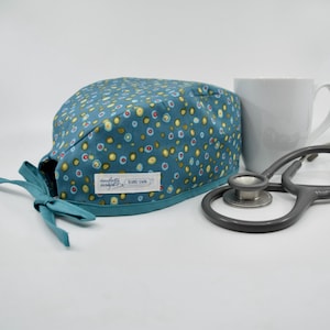 May include: Teal scrub cap with a colourful floral pattern, a teal bow, and a white label. A white mug and a grey stethoscope are next to the cap. The label reads "daughterly designs".