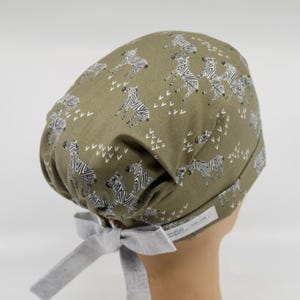 May include: Olive green scrub cap with a repeating pattern of black and white zebras and white arrow-like shapes. The cap has a light gray bow in the back for adjusting the fit. The cap is designed for medical professionals.