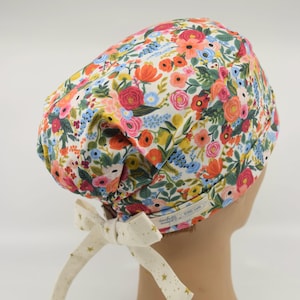 May include: A floral patterned scrub cap featuring a vibrant mix of red, orange, pink, and blue flowers on a white background. The cap has a cream-colored bow with gold stars. The cap is designed for medical professionals.