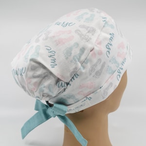 May include: White scrub cap with a pattern of baby footprints in gray, pink, and blue. The word "nurse" is written in a script font across the cap. A light blue bow is tied at the back.