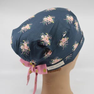 May include: A navy blue scrub cap with a floral pattern of pink and yellow flowers. The cap has a pink bow with yellow polka dots. The cap is designed to be worn during medical procedures.