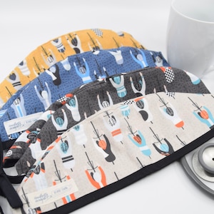 May include: A stack of four patterned scrub caps. The caps feature a repeating design of cyclists in various colors. The caps are in shades of yellow, blue, gray, and beige. Each cap has a black trim and a tag that reads "dougherty designs".