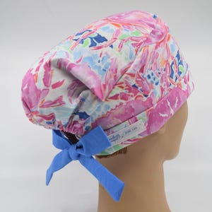 May include: A colorful scrub cap with a watercolor floral pattern in pink, blue, and green. The cap has a blue bow in the back and a white label with text. The cap is designed to be worn in a medical setting.