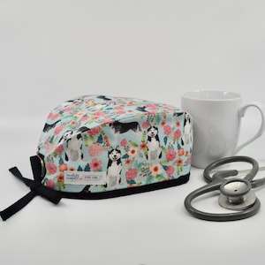 May include: A light blue surgical scrub cap with a black trim, featuring a pattern of Siberian huskies and floral elements. A white mug and a stethoscope are also in the image.