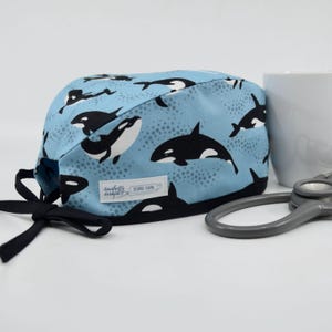 May include: A light blue surgical scrub cap featuring a pattern of black and white orca whales. The cap has a black trim and ties, with a small label that reads "Daughter Designs". A white mug and stethoscope are in the background.