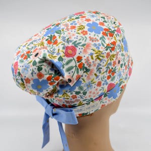 May include: A floral patterned scrub cap featuring a white background with blue, pink, and orange flowers. The cap has a light blue trim and matching ties. The design includes a variety of flowers and green leaves.