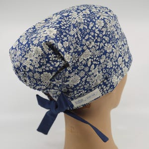 May include: A navy blue scrub cap with a white floral pattern and a matching bow. The cap is made of fabric and has a tag with text. The cap is designed to be tied at the back.