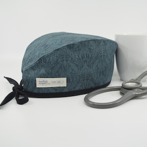 May include: A teal patterned scrub cap with a black trim and ties. The cap has a detailed floral design and a small label with text. A white mug and a gray stethoscope are in the background.