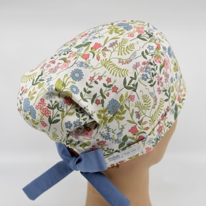 May include: A scrub cap featuring a white background with a colorful floral pattern of small blue, pink, and green flowers and leaves. The cap has a solid blue fabric tie at the back and a small white rectangular label with text on the side.