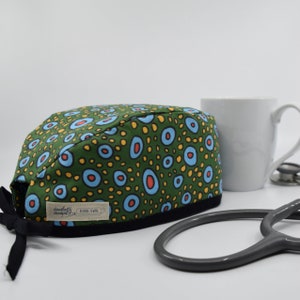 May include: A green patterned scrub cap with blue, orange, and yellow circular designs. The cap has a black brim and black ties. A white mug and a stethoscope are in the background. The cap has a tag that reads "daugherty designs bespoke sewing."