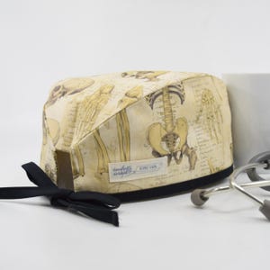 May include: A beige medical scrub cap featuring a vintage anatomical skeleton print. The cap has a black trim, a black tie, and a small label that reads "Daughters Designs". A stethoscope rests nearby.