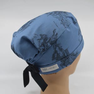 May include: A blue scrub cap featuring a dark gray sketch-style print of horses and riders. The cap has a gathered back with black fabric ties and a small white rectangular label on the side with the text "handcrafted designs" and "global sewing".