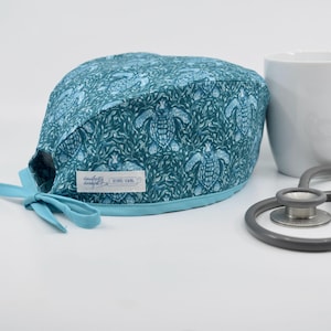 May include: A teal scrub cap with a light blue turtle and leaf pattern. The cap has a light blue trim and ties. The label reads "Daugherty Designs" and "REBEL SEWING". A white mug and stethoscope are in the background.