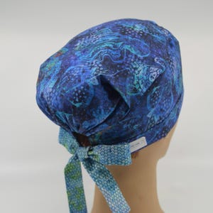 May include: A blue patterned scrub cap with a bow. The cap features a swirling design in shades of blue and teal. The bow and ties are a lighter blue with a small scale pattern. The cap is designed to be worn during medical procedures.