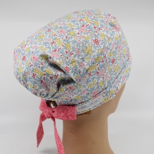 May include: A floral patterned scrub cap in a light blue, pink, and yellow color scheme. The cap features a pink bow in the back and is designed to be worn on the head. The fabric is covered in a small flower pattern.