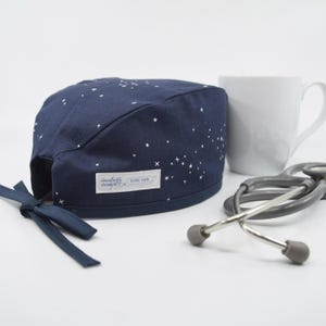 May include: A navy blue scrub cap featuring a white star pattern and a tie-back closure. A white rectangular fabric label with the text "daugherty designs" is stitched onto the side. A grey stethoscope and a white mug are positioned nearby.