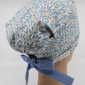 May include: A patterned surgical scrub cap featuring a botanical design with blue leaves, pink berries, and tan accents on a cream background. The cap has a denim blue bow at the back for adjustment. The cap is designed for medical professionals.