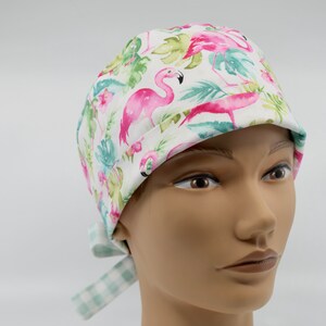 Puede incluir: Un gorro de quirófano blanco con estampado de flamencos rosas y hojas tropicales. El gorro tiene una corbata a cuadros verdes y blancos.