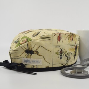 May include: A beige surgical cap featuring vintage insect illustrations. The cap has a black trim and a black tie. A white label with text is visible. A stethoscope and a white mug are in the background.